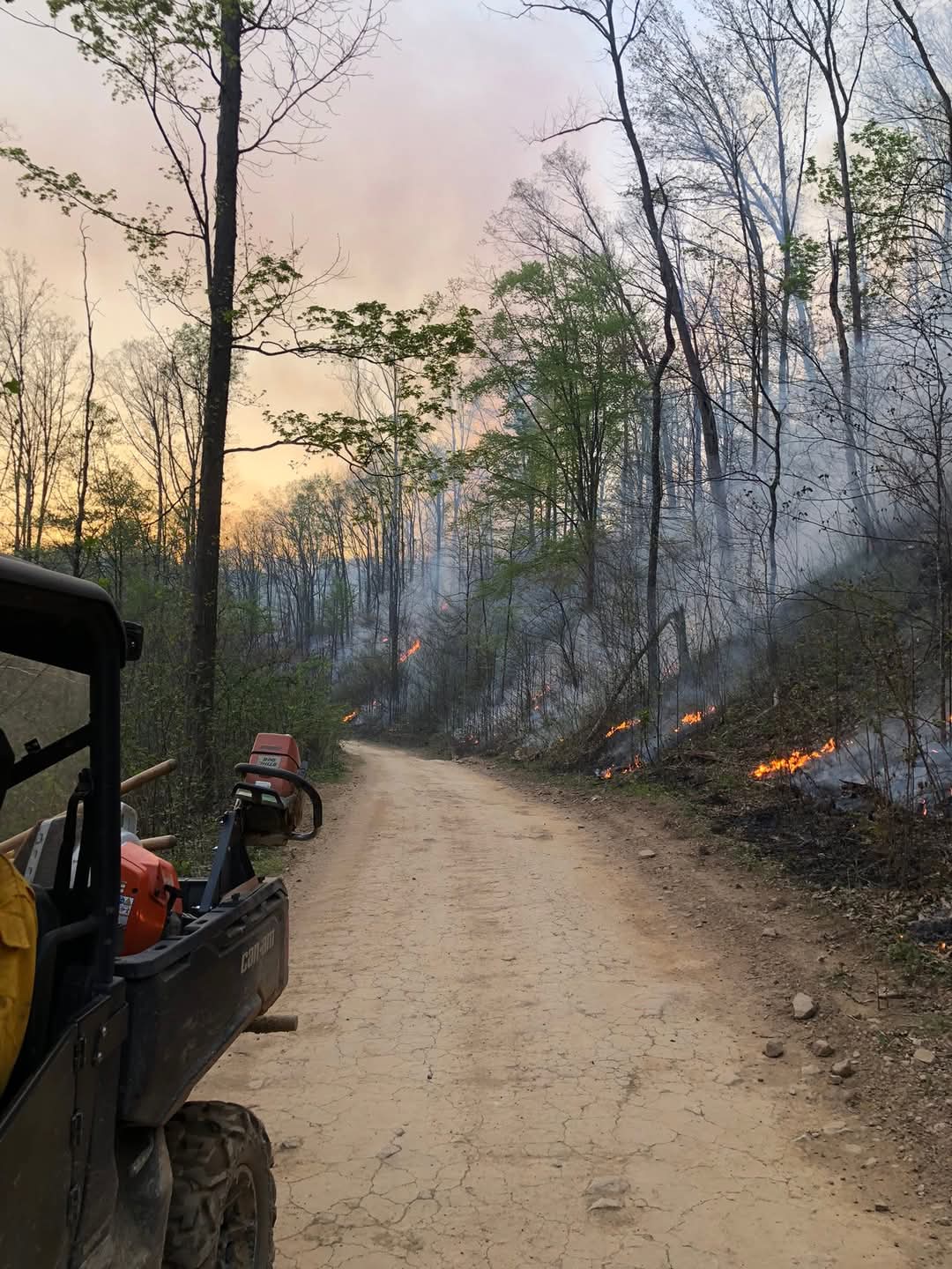 A UTV on a dirt road across from fire on a hill.